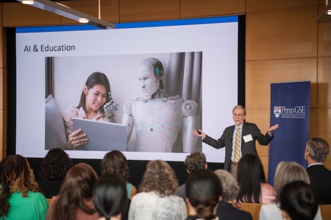 Dr. Lester gives a lecture in front of a packed crowd in front of a screen with an image of  a woman and a robot sharing a laptop and the words "AI in Education" written above it. 