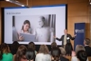 Dr. Lester gives a lecture in front of a packed crowd in front of a screen with an image of  a woman and a robot sharing a laptop and the words "AI in Education" written above it. 