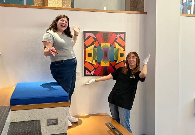 Two young women pose in front of a multicolored abstract art piece along the staircase in the Penn GSE lobby.