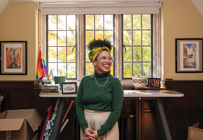 Krista Cortes stands, hands folded in front of her, laughing in the middle of her office surrounded by decorations showcasing Latin American culture.
