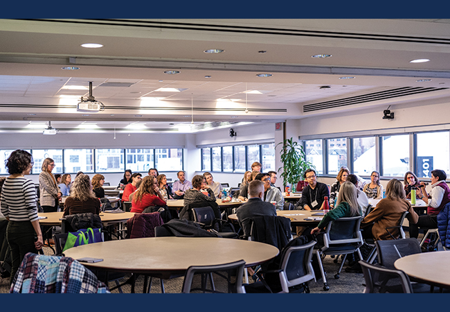 A group of more than 30 teacher education researchers gathered around large round tables in a conference room listen to one of the attendees speaking into a microphone while the presenters stand at the front.