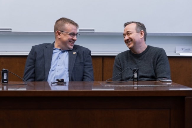 Representatives Bryan Cutler and Peter Schweyer sit at a table in front of a white board and face each other, laughing.