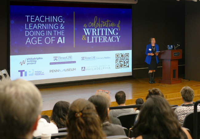Amy Stornaiuolo speaks into a microphone on stage in front of an audience at the 2024 Celebration of Writing and Literacy with the conference logo on a screen behind her.