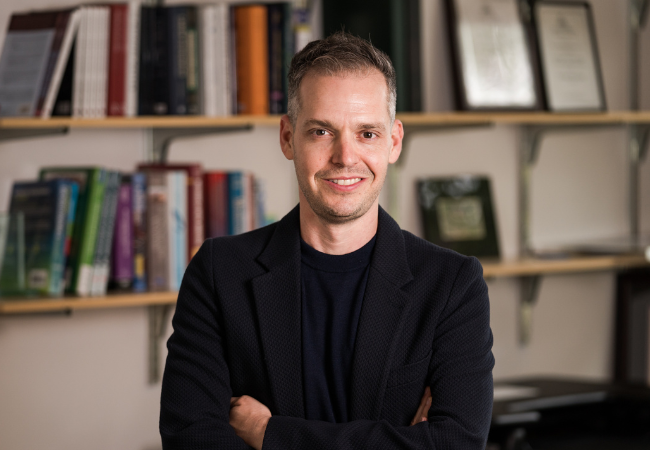 Portrait of Michael Gottfried, standing with arms crossed, smiling, with bookshelves in the background.