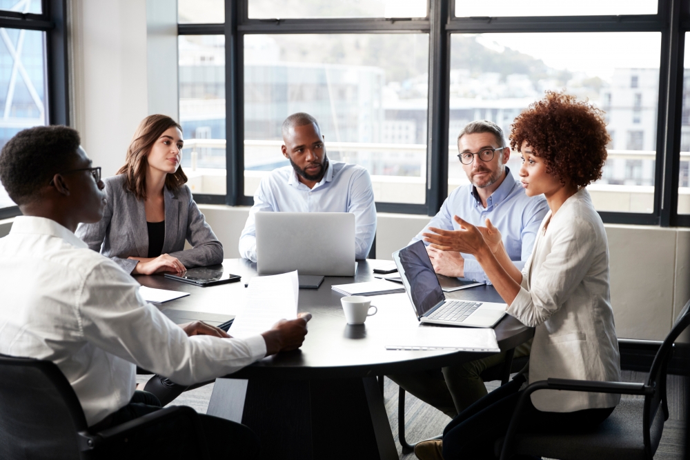 Four people sit around a table listening to a fifth person speak.