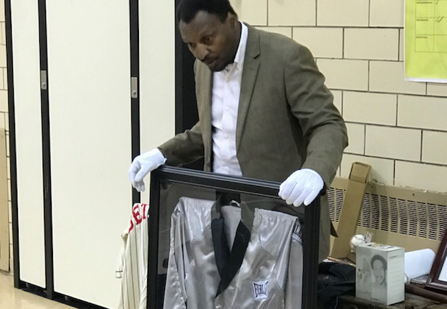 Randy Hayward holds up a frame containing a boxing robe of Muhammad Ali using white gloves to share it with a group of students. 