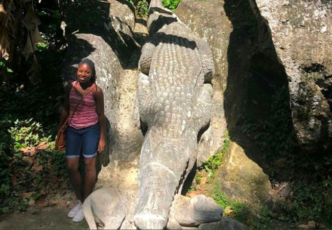 Angela Crumdy stands beside a large statue of an alligator biting another animal, surrounded by trees and rocks in Guantánamo, Cuba.