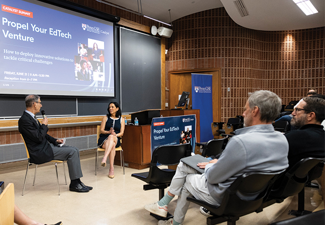 Penn GSE Dean Katharine Strunk, seated in a chair in front of an audience in a lecture hall, speaks into a microphone as Vice Dean of Innovative Programs and Partnerships Michael Golden listens, seated in a chair next to her. A screen behind them reads "Catalyst Summit: Propel Your EdTech Venture | How to deploy innovative solutions to tackle critical challenges."