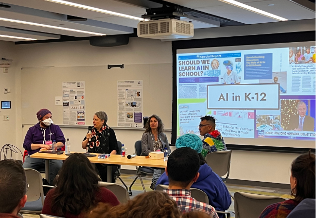 Four people sit in a row at a long table in front of an audience with a slide that says “AI in K–12” on a screen behind them.