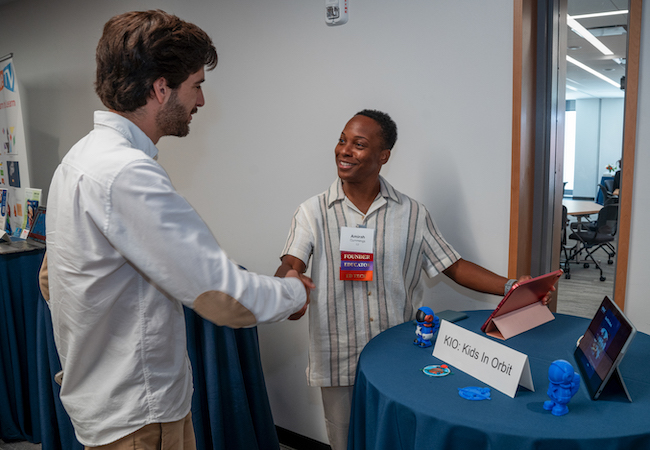An alumni founder shakes the hand of another alum in front of the table for his venture, KIO: Kids in Orbit, which features iPads and small spaceman figurines