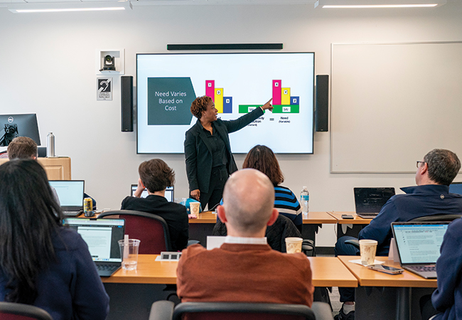 Adult students seated at rows of tables in the foreground facing foreward as an instructor standing in front of them points to a colorful financial aid graph on a screen titled “Need Varies Based on Cost.”