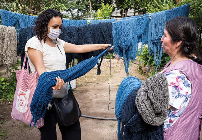 Fabiola Lara stands wearing a medical mask with a woman artisan outdoors in Oaxaca, Mexico, holding and examining a pile of threads, with more threads hanging behind them.