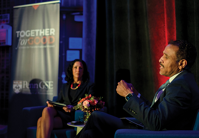 Superintendent Watlington speaks, seated in the foreground, as Dean Strunk, seated in the background in front of a “Together for Good: Penn GSE” banner, listens.