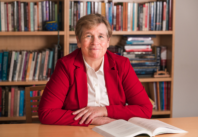 Karen Weaver smiles, wearing a red blazer and seated at a desk in front a full bookshelf