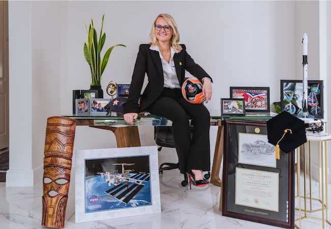 Lauren in a suit sitting on a desk surrounded by awards, framed diplomas, a model rocket, plants, space images, and sports memorabilia.