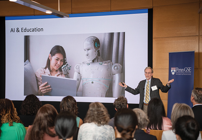 A speaker delivers a talk about AI & education in front of a screen featuring a robot teaching a student on an iPad