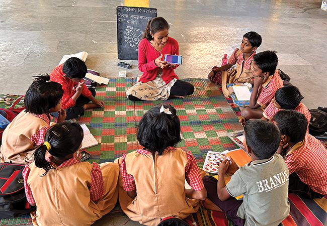 A teacher displays a chatbot on her phone screen while sitting cross-legged on a mat on the floor alongside a circle of her students during a Saturday Art Class in India