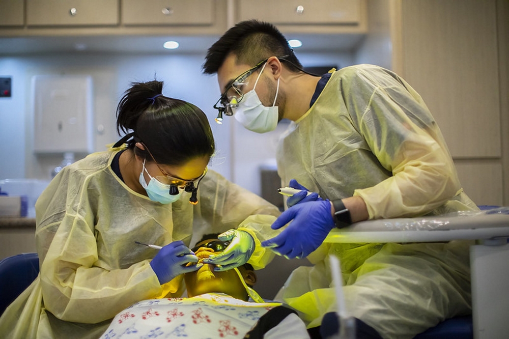 Two health students in masks, glasses, and yellow scrubs work over a patient.