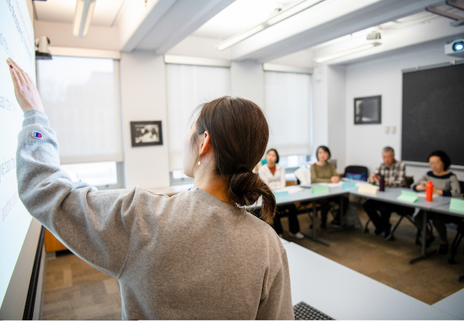 A GSE student points to her lesson on the board and she instructs a small classroom of adult learners who are seated around a table. 