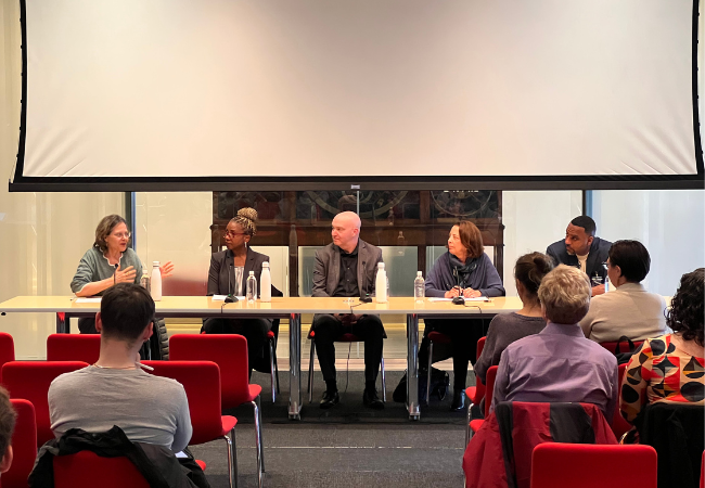 Five professors sit behind a table speaking with one another in front of an audience.