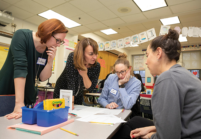 RMT trainers stand over a table and instruct two teachers, seated, in the teaching method