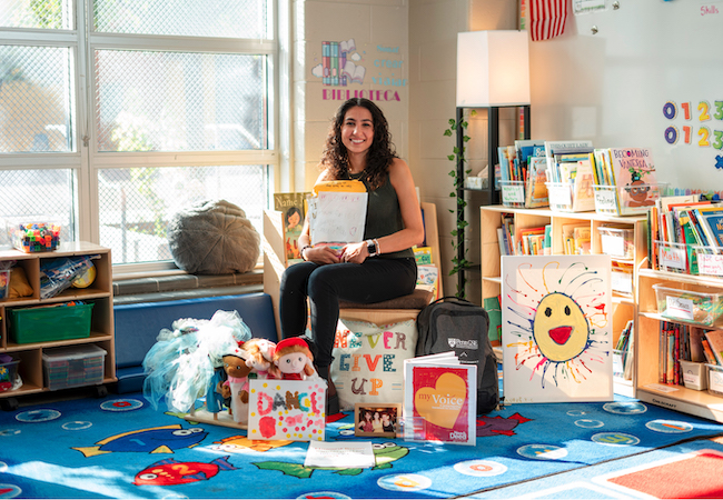 Sarah sits in her classroom holding a packet of letters and drawings from students and surrounded by puppets, books, pillows, and costumes that she uses in her class.