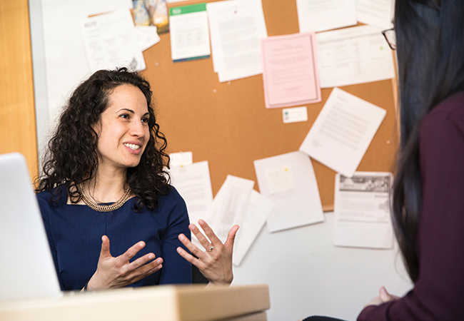 Sharon Wolf seated at her desk, smiling and gesturing with her hands as she speaks to a student