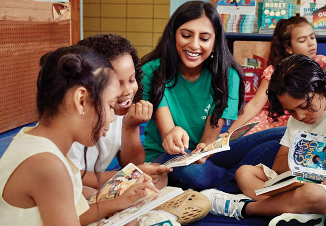 Penn GSE alum Rina Madhani sits cross-legged on the floor surrounded by elementary schoolchildren, all reading books and laughing together