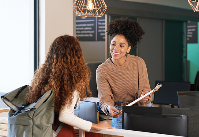 A smiling counselor leans over a counter with a clipboard while helping a teen with a backpack