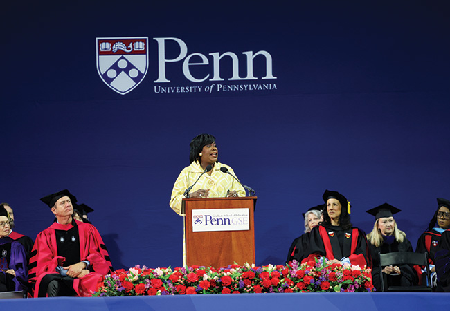 Mayor Cherelle Parker speaks at a podium while Dean Katharine O. Strunk and other faculty in regalia look on at Penn GSE's 2024 commencement ceremony