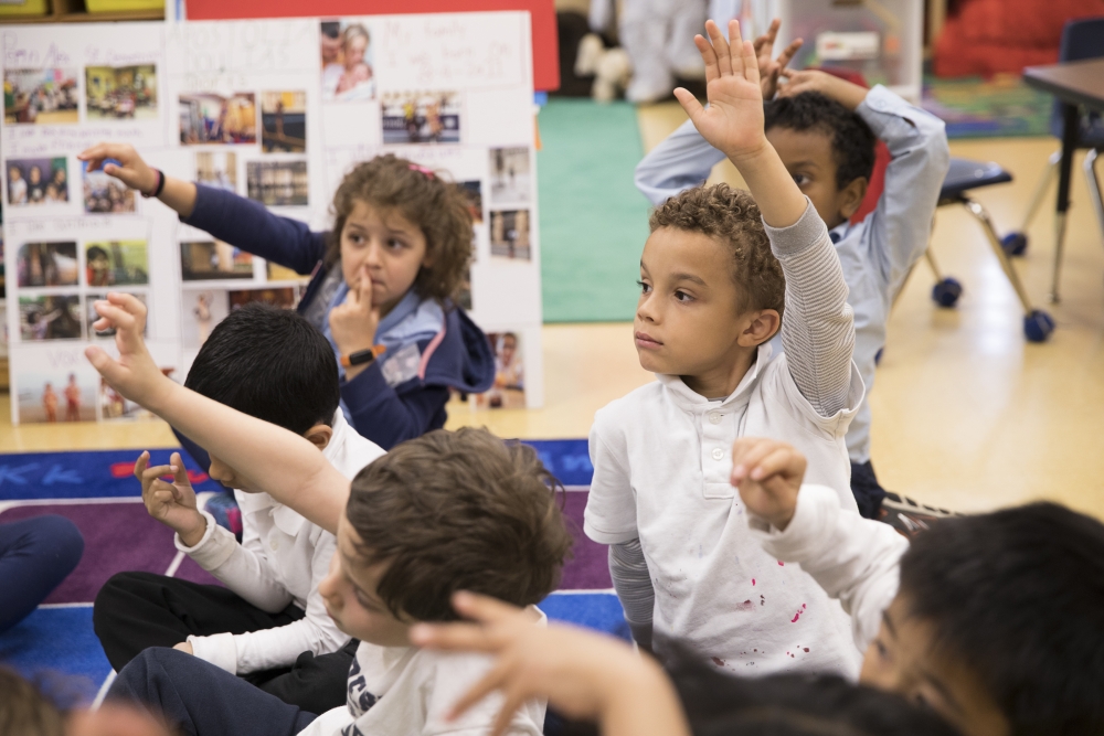 Students sitting on a mat raising their hands for the teacher