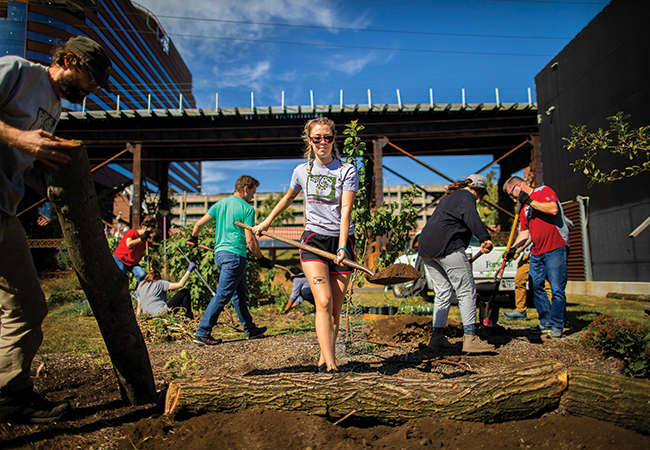 A young woman shoveling dirt at the Penn Park Orchard with other volunteers working and Patient Pavilion in the background