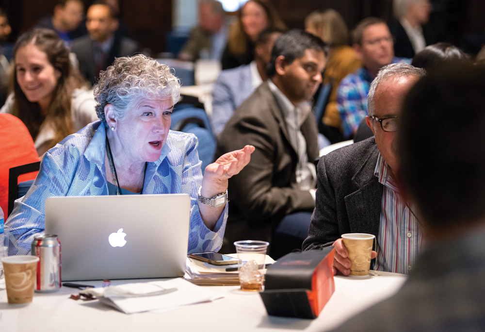 People engaging in conversation at a conference, with a woman speaking and gesturing near a laptop.