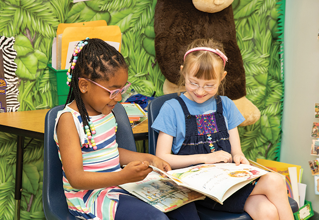 Two school girls sitting next to each other on blue chairs are smiling and flipping through pages of a large book with colorful illustrations.