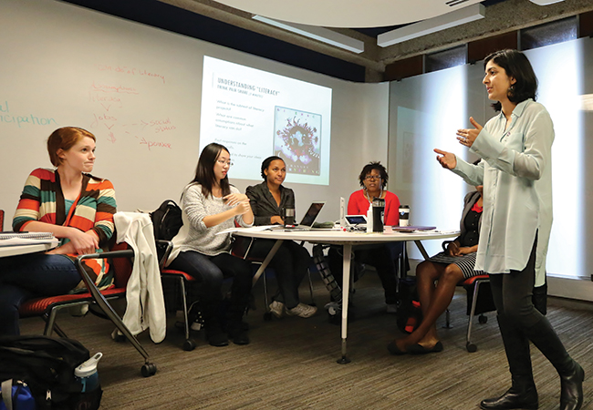 A lecturer wearing a light green colored blouse addresses a classroom of diverse students seated around round tables. The title of the digital presentation in the background reads “Understanding “Literacy”