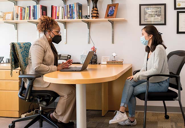 Dr. Andrea Kane is seen wearing a mask and speaking to a student sitting opposite to her in an office.