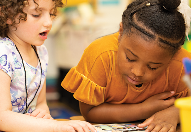 Close-up image of two children at a desk looking down to read side by side