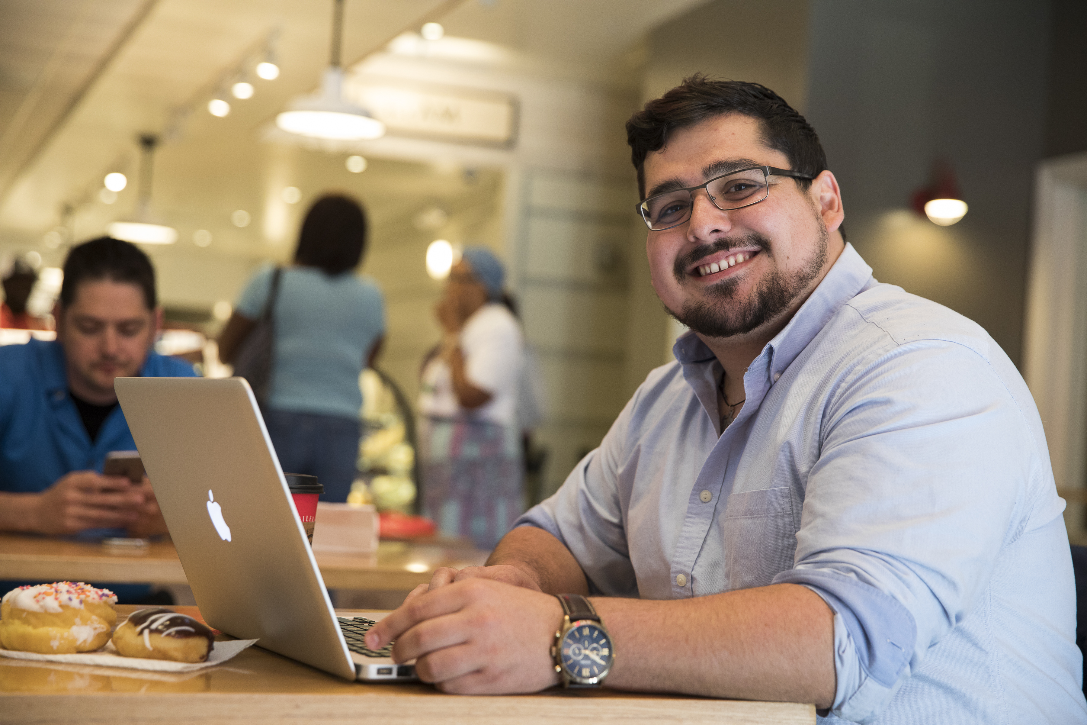 A graduate student smiles at the camera, sitting and working at his laptop