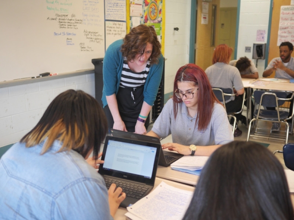 A student-teacher, standing, working with three seated high school students.