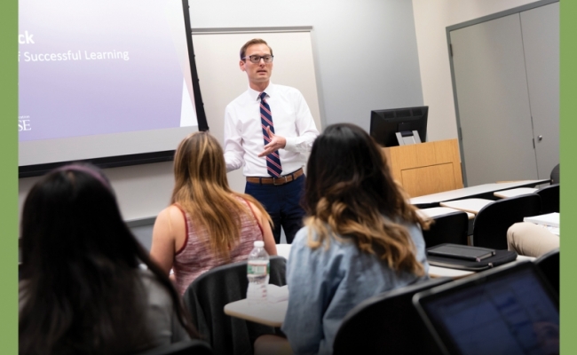 A man wearing glasses and professional attire stands and teaches in front of a classroom.