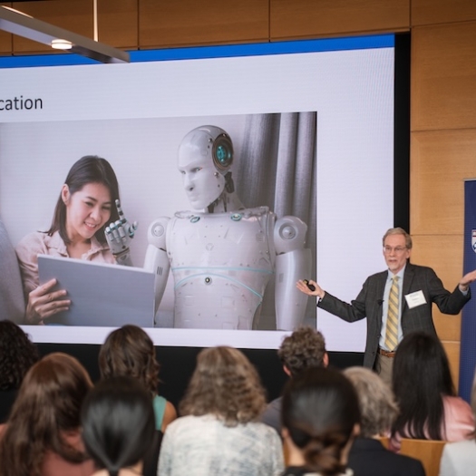 Dr. Lester gives a lecture in front of a packed crowd in front of a screen with an image of  a woman and a robot sharing a laptop and the words "AI in Education" written above it. 