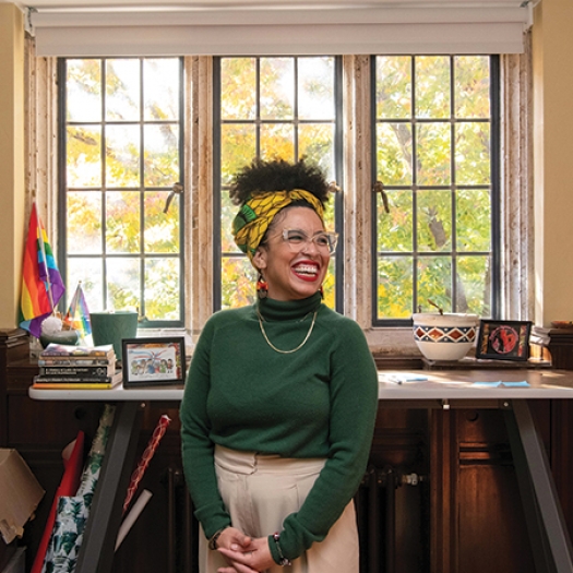 Krista Cortes stands, hands folded in front of her, laughing in the middle of her office surrounded by decorations showcasing Latin American culture.