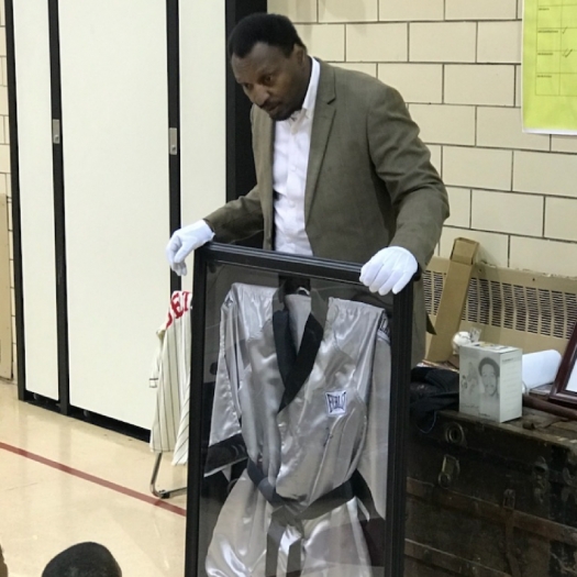 Randy Hayward holds up a frame containing a boxing robe of Muhammad Ali using white gloves to share it with a group of students. 