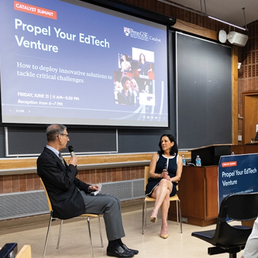 Penn GSE Dean Katharine Strunk, seated in a chair in front of an audience in a lecture hall, speaks into a microphone as Vice Dean of Innovative Programs and Partnerships Michael Golden listens, seated in a chair next to her. A screen behind them reads "Catalyst Summit: Propel Your EdTech Venture | How to deploy innovative solutions to tackle critical challenges."