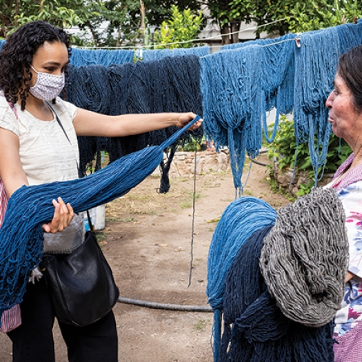 Fabiola Lara stands wearing a medical mask with a woman artisan outdoors in Oaxaca, Mexico, holding and examining a pile of threads, with more threads hanging behind them.