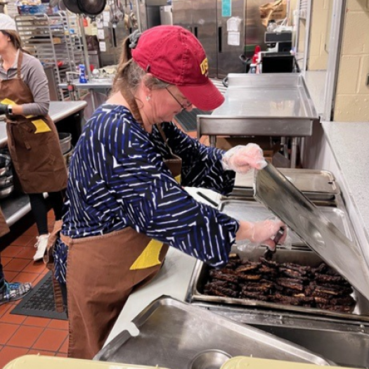 Julee Gard, wearing a red cap, a navy blue and white striped shirt, and a brown apron, is focused on serving sausages at a soup kitchen in Joliet, Illinois, alongside her University of St. Francis colleagues.