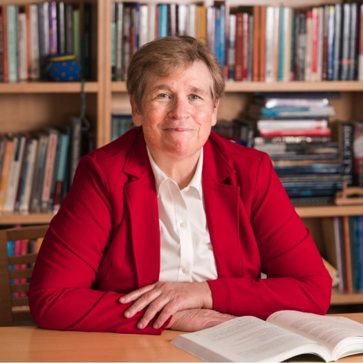 Karen Weaver smiles, wearing a red blazer and seated at a desk in front a full bookshelf