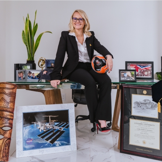 Lauren in a suit sitting on a desk surrounded by awards, framed diplomas, a model rocket, plants, space images, and sports memorabilia.