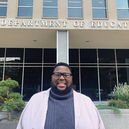 Mark Ziegler-Thayer smiles at the camera wearing a white blazer over a gray sweater, standing in front of the Lyndon Baines Johnson Department of Education Building in Washington, D.C. The words “U.S. Department of Education” are mounted on the face of the building in large metal letters above Mark’s head.