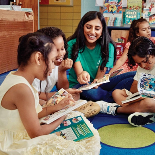 Penn GSE alum Rina Madhani sits cross-legged on the floor surrounded by elementary schoolchildren, all reading books and laughing together